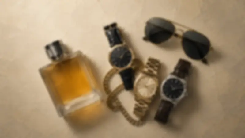 Aviator sunglasses, amber fragrance bottle, three watches, and gold chain on textured cream plaster.