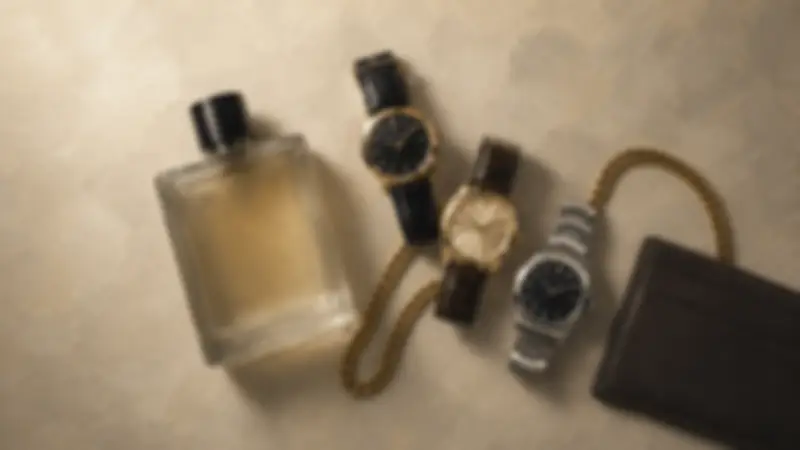 Black wallet, amber fragrance, three classic watches, and a gold chain arranged on pale plaster.
